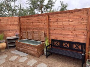 Detaljebillede af et udendørs koldvandsbad i trækasse, placeret ved siden af en sort bænk og et højt træhegn.